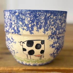 May include: A ceramic bowl with a blue speckled rim and a cream-colored body. The bowl features a hand-drawn cow illustration with black spots and a pink udder. The cow stands on a green grassy area.