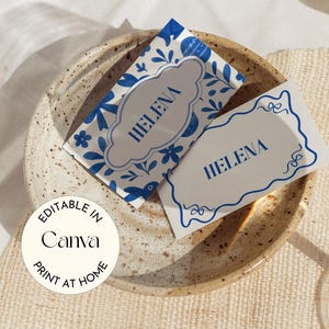 Könnte beinhalten: Zwei Tischkarten mit dem Namen "HELENA" in Blau, eine mit floralem Design. Die Karten liegen auf einem gesprenkelten Keramikteller. Eine halbe Zitrone und ein Glas Wasser sind ebenfalls im Bild. Der Text auf dem Bild lautet "EDITABLE IN Canva PRINT AT HOME."
