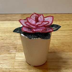 May include: A handcrafted beaded flower in a white ceramic pot. The flower has pink and red petals with a beaded green leaf base. The pot has a ribbed texture. The flower is a decorative item.