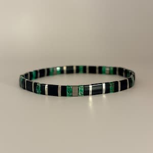 Peut inclure: Un bracelet noir et vert avec des accents argentés. Le bracelet est composé de perles rectangulaires noires et vertes, séparées par des barres métalliques argentées. Le bracelet est de style extensible et est présenté sur un fond neutre.