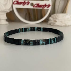 Peut inclure: Un bracelet noir avec des perles rectangulaires turquoise et noires. Le bracelet porte le texte "Charley's" dans un cercle blanc en arrière-plan. Un accessoire élégant.