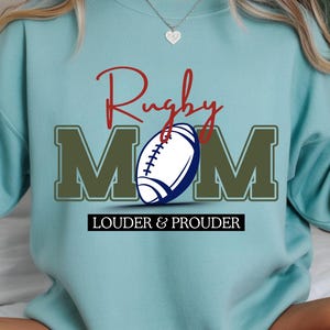 Può includere: Felpa azzurra con la scritta "Rugby Mom" in corsivo rosso e lettere verdi. Un pallone da rugby bianco e blu è al centro. Il testo "LOUDER & PROUDER" è in basso.