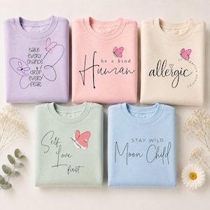 Può includere: Cinque felpe color pastello con citazioni ispiratrici e grafiche di farfalle. Le magliette sono lavanda, rosa, crema, verde menta e azzurro. Il testo include "take every chance drop every fear", "be a kind human", "allergic to negative energy", "self love first" e "stay wild moon child".