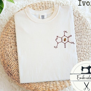Könnte beinhalten: Elfenbeinfarbenes T-Shirt mit einem gestickten Koffeinmolekül-Design in Braun. Das Shirt ist auf einer gewebten, kreisförmigen Matte gefaltet. Das Wort "Ivory" ist über dem Shirt aufgedruckt. Ein Logo mit der Aufschrift "Embroidered" befindet sich unten rechts.