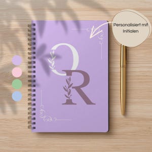Könnte beinhalten: Ein lila Spiralnotizbuch mit den Buchstaben "OR" in Weiß und Lila, verziert mit floralen Akzenten. Ein goldener Stift liegt neben dem Notizbuch. Der Text "Personalisiert mit Initialen" ist sichtbar.