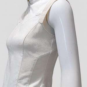 May include: A sleeveless white top featuring a high mock neck collar with a front zipper closure. The garment is constructed with structured vertical paneling and wide shoulder straps, paired with a metallic silver belt at the waistline.