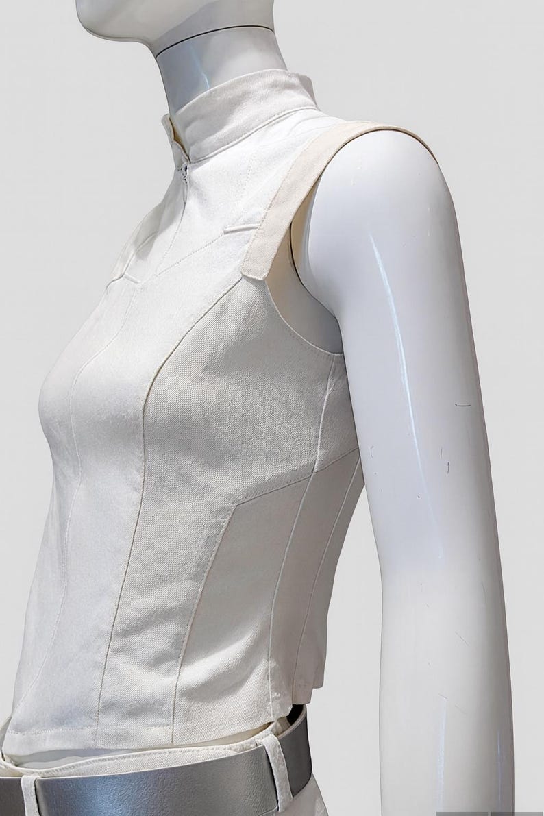 May include: A sleeveless white top featuring a high mock neck collar with a front zipper closure. The garment is constructed with structured vertical paneling and wide shoulder straps, paired with a metallic silver belt at the waistline.
