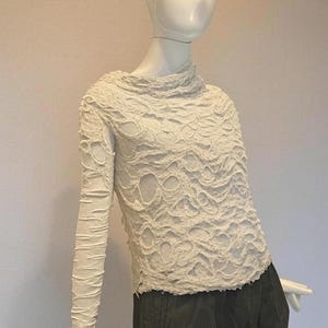 May include: A long-sleeved off-white top featuring a heavily textured, distressed surface with numerous irregular cutouts and frayed edges. The garment has a high, draped neckline and a fitted silhouette, worn over dark olive green trousers.