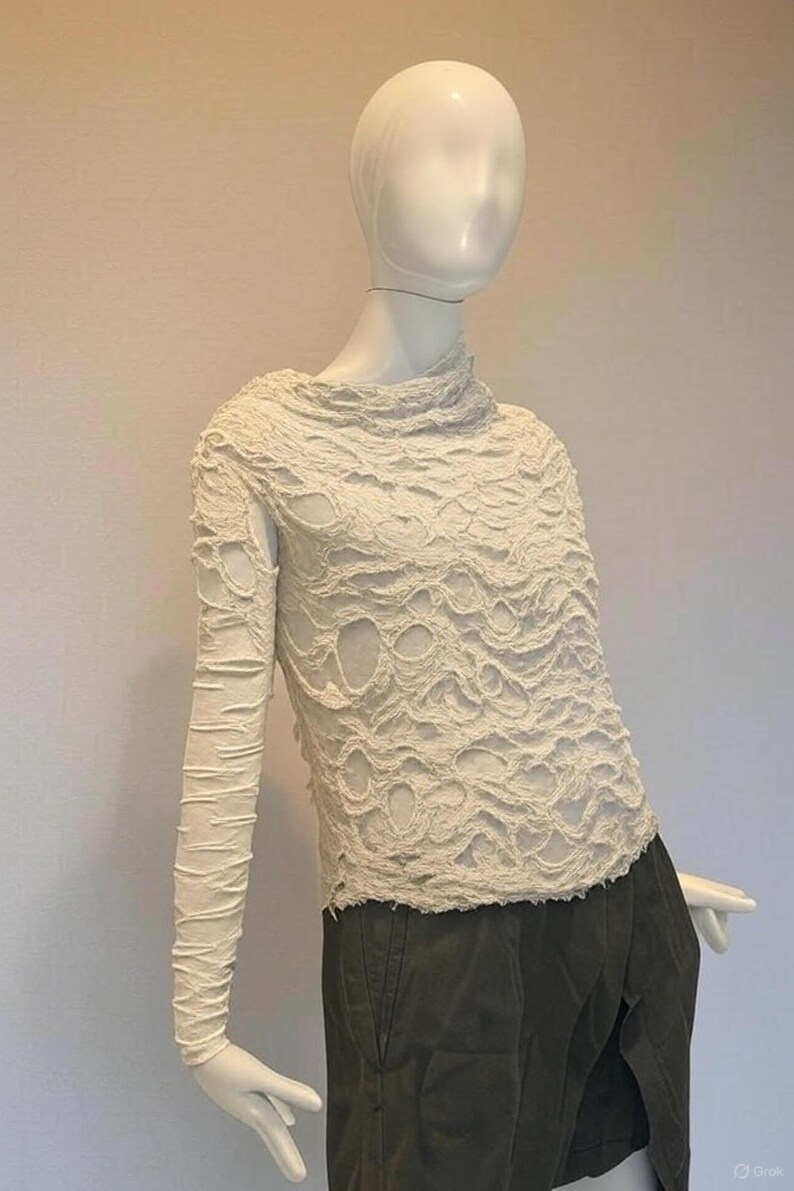 May include: A long-sleeved off-white top featuring a heavily textured, distressed surface with numerous irregular cutouts and frayed edges. The garment has a high, draped neckline and a fitted silhouette, worn over dark olive green trousers.