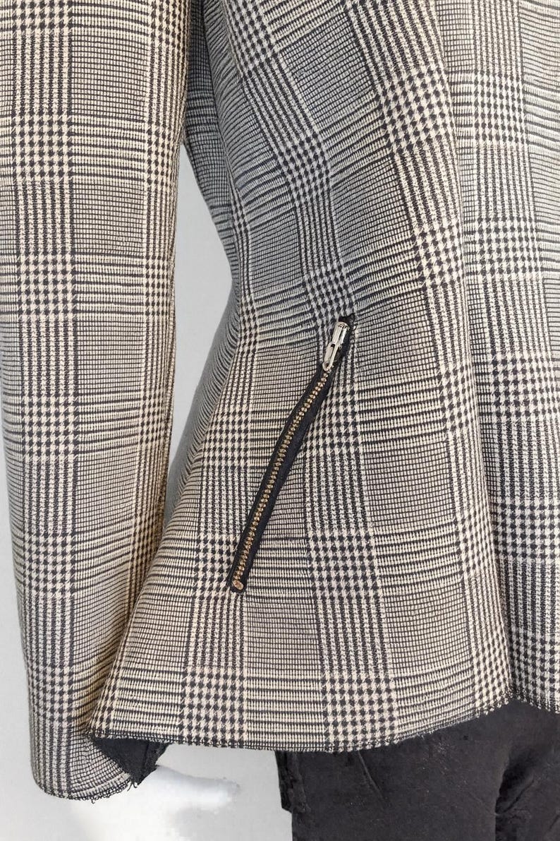 May include: A close-up of a black and white plaid blazer. The jacket features a diagonal zipper pocket and a black lining. The fabric has a classic glen plaid pattern.