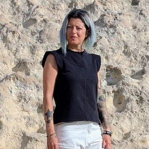 May include: A black sleeveless top with structured shoulders and visible seam detailing across the chest, paired with white pants. The top features a round neckline and a straight hemline, constructed from a solid black fabric with distinct stitching.