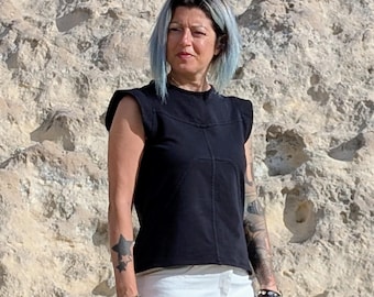 May include: A black sleeveless top with structured shoulders and visible seam detailing across the chest, paired with white pants. The top features a round neckline and a straight hemline, constructed from a solid black fabric with distinct stitching.