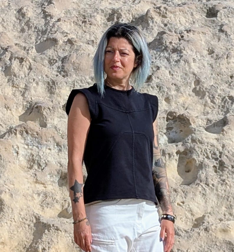 May include: A black sleeveless top with structured shoulders and visible seam detailing across the chest, paired with white pants. The top features a round neckline and a straight hemline, constructed from a solid black fabric with distinct stitching.