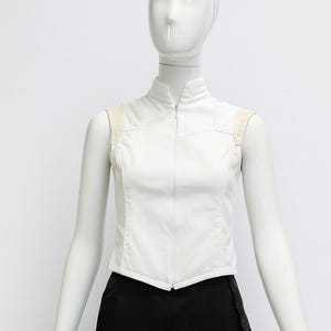 May include: A sleeveless white vest featuring a high collar and a full-length front zipper closure. The garment includes light beige shoulder panels and a structured, fitted silhouette that tapers at the waist with a pointed hemline.