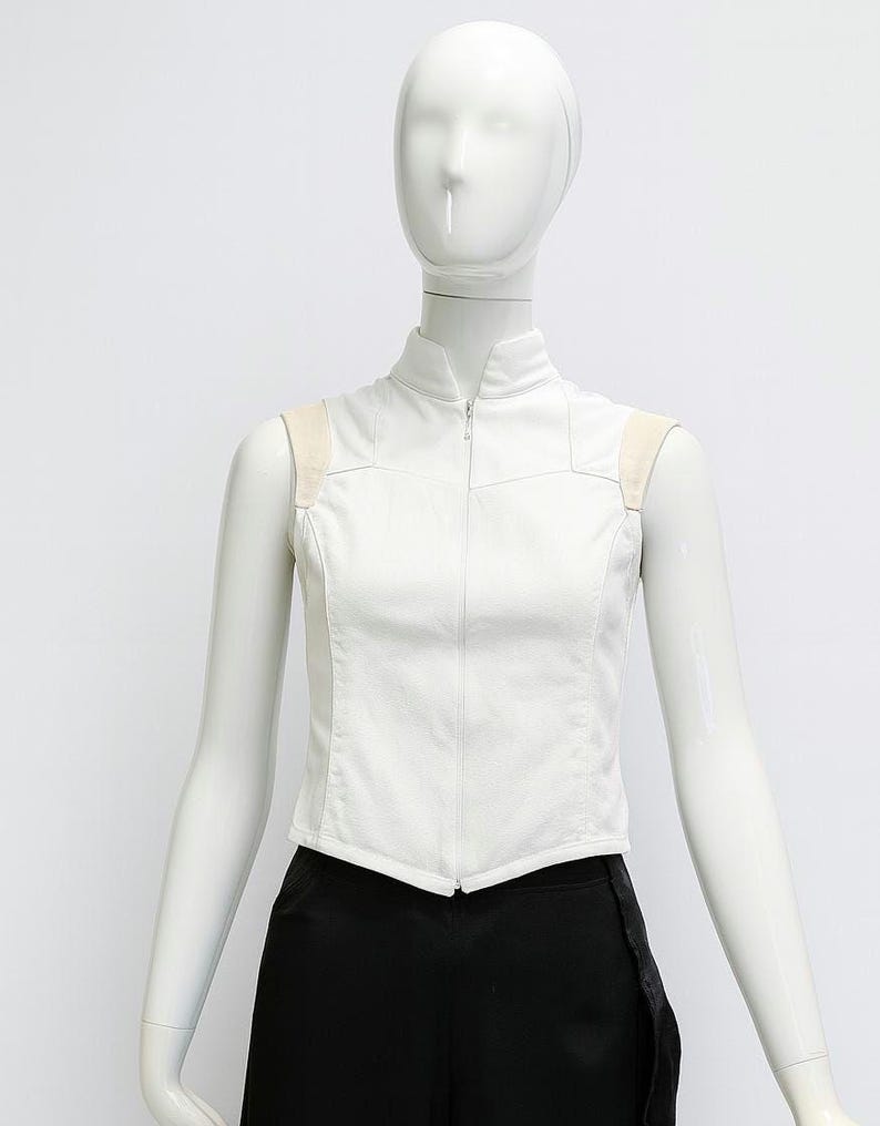 May include: A sleeveless white vest featuring a high collar and a full-length front zipper closure. The garment includes light beige shoulder panels and a structured, fitted silhouette that tapers at the waist with a pointed hemline.