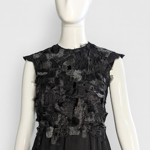Puede incluir: Un vestido negro sin mangas con un corpi&ntilde;o texturizado compuesto por parches de tela deshilachados y superpuestos en varios tonos de negro y gris oscuro. La parte inferior del vestido consiste en una falda de tela negra lisa.