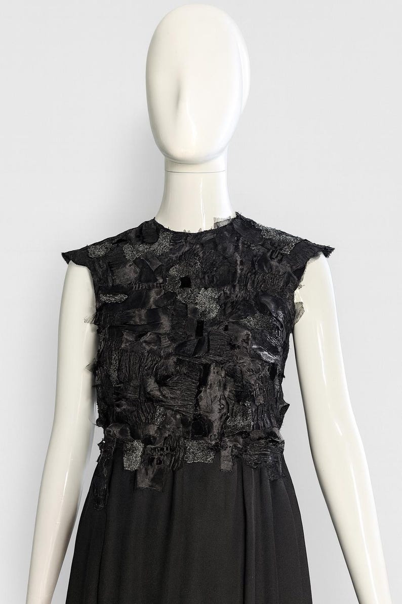 Puede incluir: Un vestido negro sin mangas con un corpi&ntilde;o texturizado compuesto por parches de tela deshilachados y superpuestos en varios tonos de negro y gris oscuro. La parte inferior del vestido consiste en una falda de tela negra lisa.