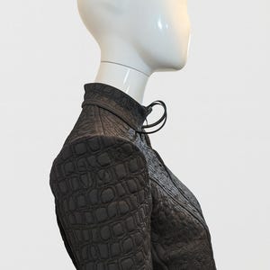 May include: A dark brown jacket with a high collar and a textured, embossed crocodile-skin pattern. The garment features long sleeves, a structured shoulder design, and a front closure with a small loop detail at the neckline.
