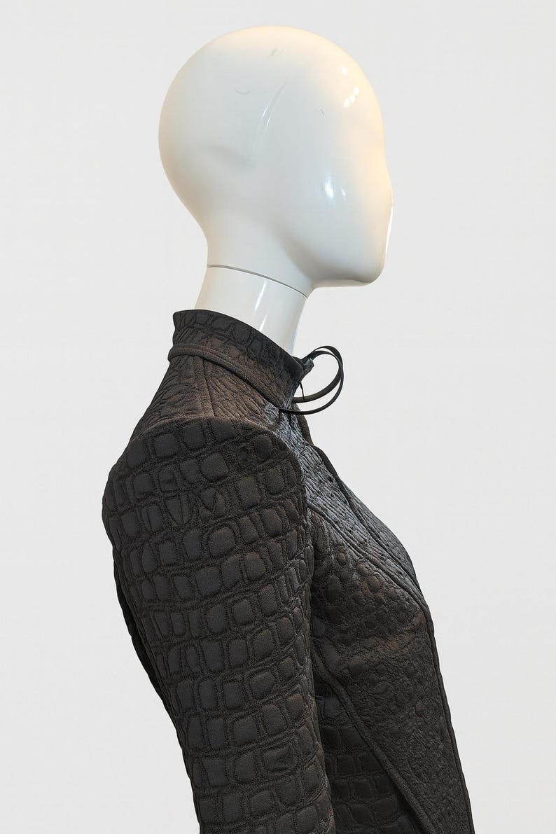 May include: A dark brown jacket with a high collar and a textured, embossed crocodile-skin pattern. The garment features long sleeves, a structured shoulder design, and a front closure with a small loop detail at the neckline.