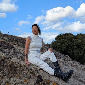 K&ouml;nnte beinhalten: Ein &auml;rmelloser wei&szlig;er Jumpsuit mit Stehkragen, einem silbernen G&uuml;rtel mit rechteckiger Schnalle und passender wei&szlig;er Hose. Das Outfit wird mit schwarzen wadenhohen Stiefeln kombiniert, die mehrere horizontale Riemen und Silberschnallen haben.