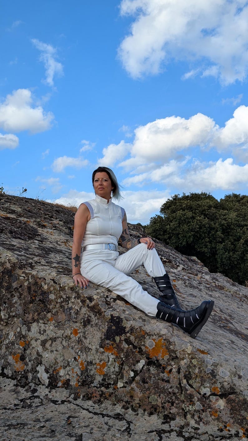 K&ouml;nnte beinhalten: Ein &auml;rmelloser wei&szlig;er Jumpsuit mit Stehkragen, einem silbernen G&uuml;rtel mit rechteckiger Schnalle und passender wei&szlig;er Hose. Das Outfit wird mit schwarzen wadenhohen Stiefeln kombiniert, die mehrere horizontale Riemen und Silberschnallen haben.