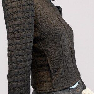 May include: A dark brown jacket with a textured, embossed crocodile-skin pattern, featuring a high collar, structured shoulders, and long sleeves. The jacket is worn over dark grey pants with a visible button at the waistline.