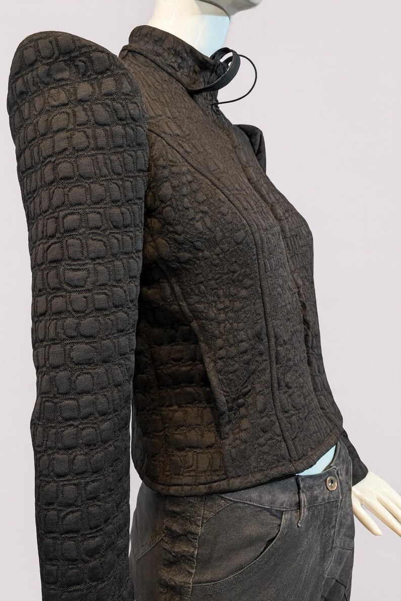 May include: A dark brown jacket with a textured, embossed crocodile-skin pattern, featuring a high collar, structured shoulders, and long sleeves. The jacket is worn over dark grey pants with a visible button at the waistline.