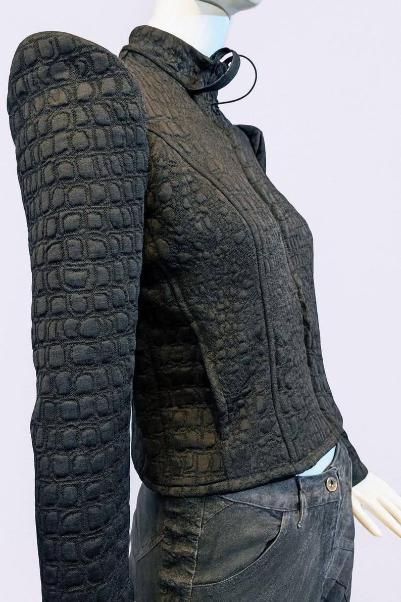 May include: A black jacket with a textured, embossed crocodile-skin pattern, featuring a high collar, structured shoulders, and long sleeves. The jacket is worn over dark gray pants with a visible button closure at the waistline.