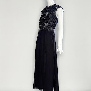Puede incluir: Un vestido negro sin mangas hasta el suelo con un corpi&ntilde;o texturizado de volantes de tela superpuestos y una falda fluida. La prenda tiene un cuello alto y una silueta recta, exhibida en un maniqu&iacute; blanco sobre un fondo liso.