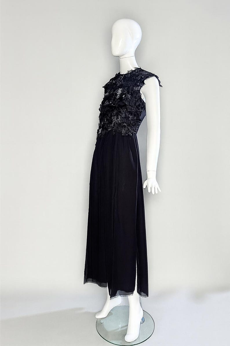 Puede incluir: Un vestido negro sin mangas hasta el suelo con un corpi&ntilde;o texturizado de volantes de tela superpuestos y una falda fluida. La prenda tiene un cuello alto y una silueta recta, exhibida en un maniqu&iacute; blanco sobre un fondo liso.