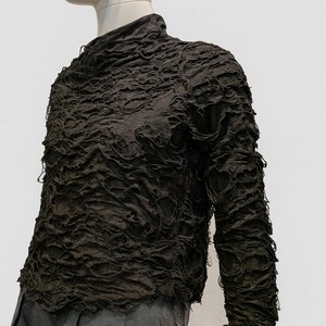 May include: A dark brown long-sleeve top with a high neckline and a heavily textured, distressed surface featuring numerous small, irregular holes and frayed edges throughout the fabric, worn over dark gray trousers.
