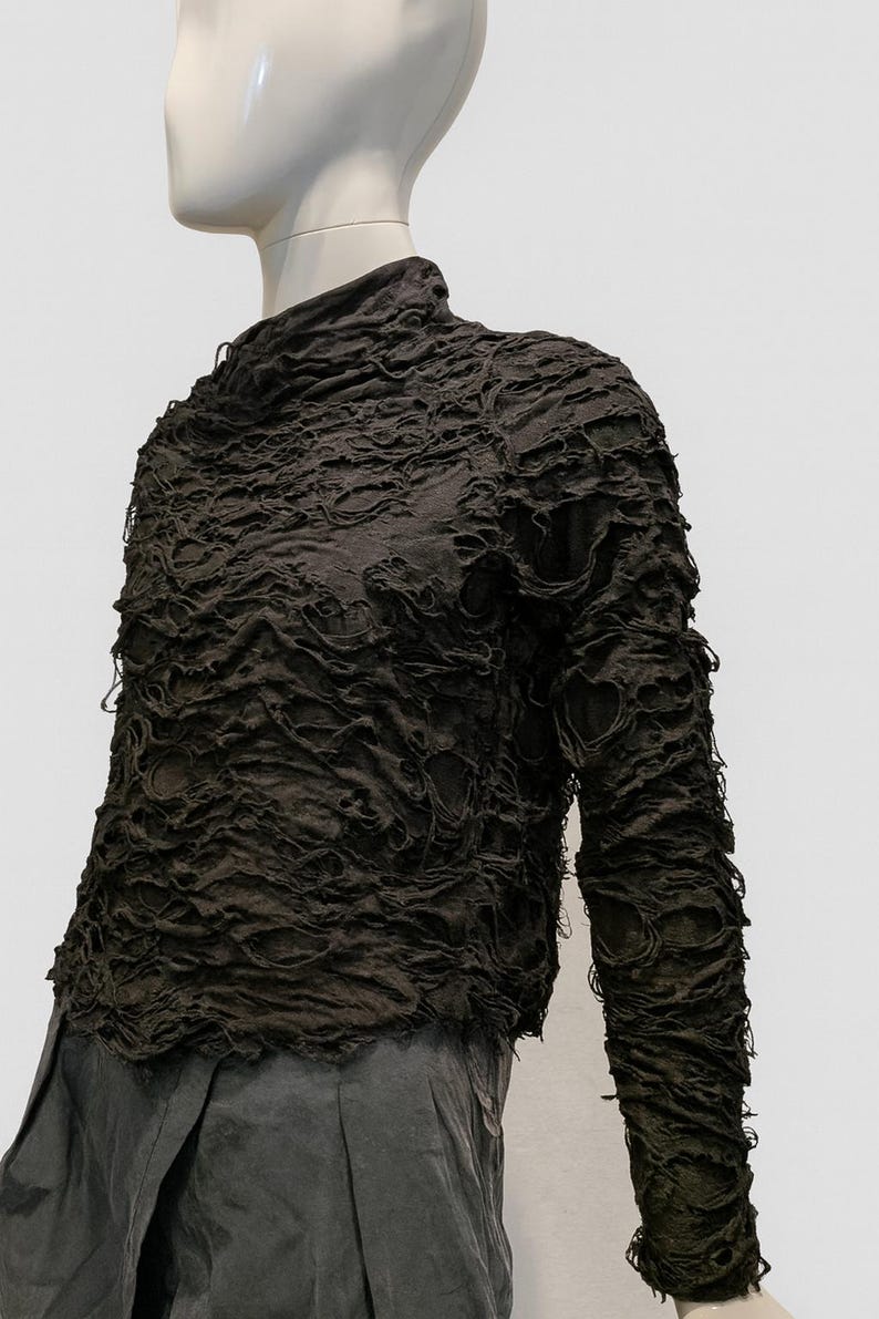 May include: A dark brown long-sleeve top with a high neckline and a heavily textured, distressed surface featuring numerous small, irregular holes and frayed edges throughout the fabric, worn over dark gray trousers.