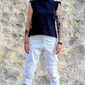 May include: A black sleeveless top with structured shoulders and a center seam, paired with white cargo pants featuring side pockets and knee detailing, worn with black mid-calf boots that have multiple horizontal straps and buckles.