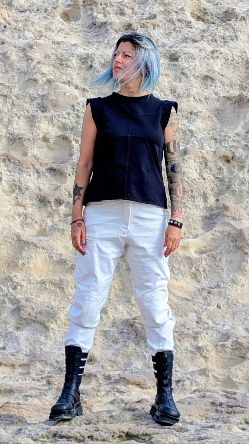 May include: A black sleeveless top with structured shoulders and a center seam, paired with white cargo pants featuring side pockets and knee detailing, worn with black mid-calf boots that have multiple horizontal straps and buckles.