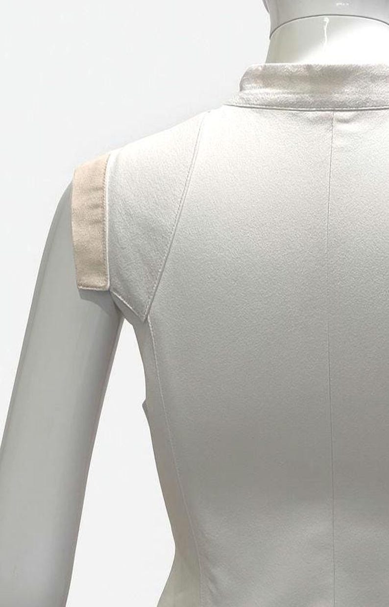 May include: A sleeveless white top featuring a high collar and a structured shoulder design with contrasting beige fabric panels. The garment is fitted, showcasing vertical seam detailing along the torso and a clean, minimalist construction.