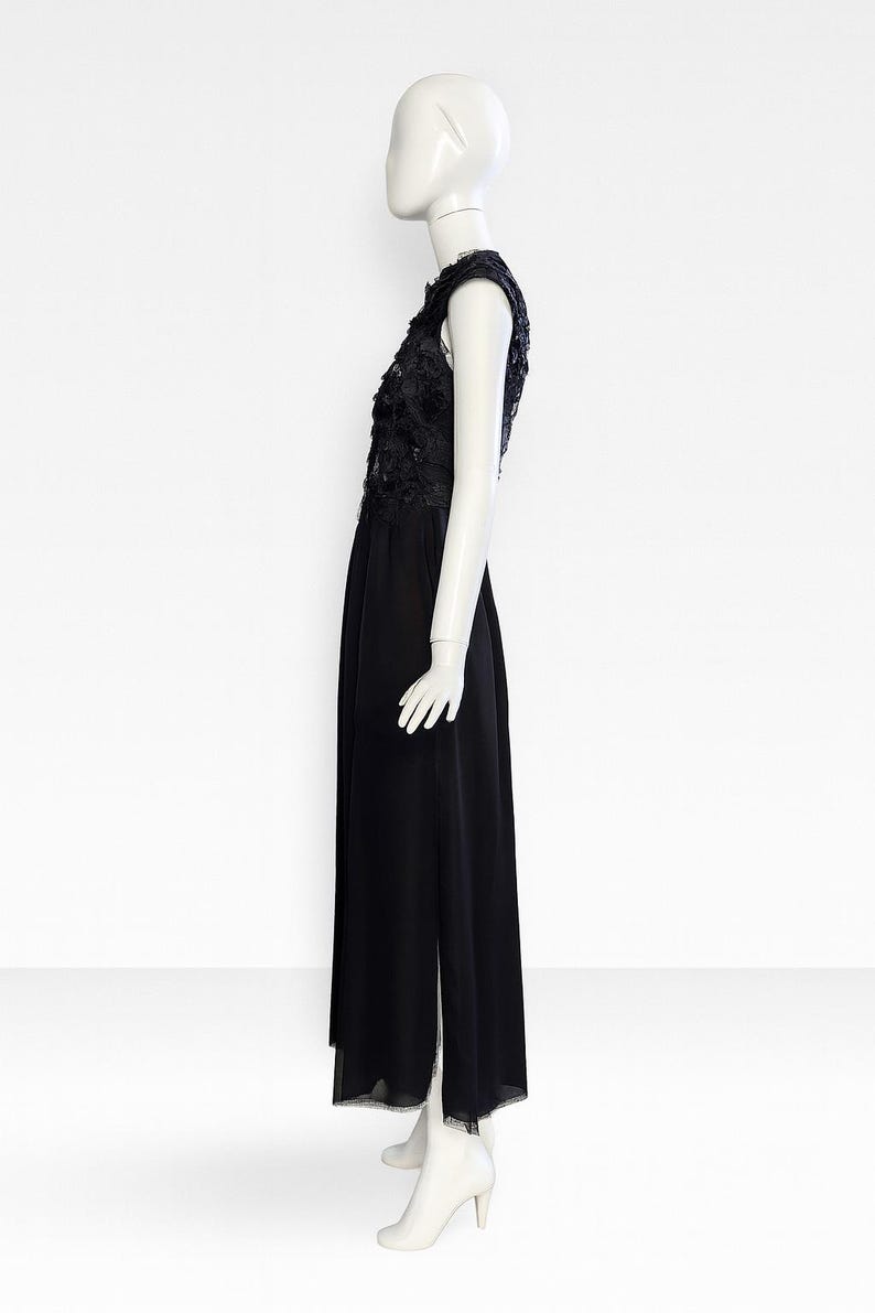 Puede incluir: Un vestido negro sin mangas hasta el suelo con un corpi&ntilde;o de encaje floral y una capa superpuesta transparente. La falda est&aacute; hecha de tela negra s&oacute;lida con una abertura lateral, exhibido sobre un maniqu&iacute; de color blanco.
