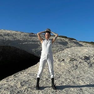 K&ouml;nnte beinhalten: Ein &auml;rmelloser wei&szlig;er Jumpsuit mit Frontrei&szlig;verschluss und Stehkragen, kombiniert mit schwarzen wadenhohen Stiefeln. Der Jumpsuit hat ein k&ouml;rperbetontes Oberteil und eine gerade geschnittene Hose, was einen monochromen Look vor einer Felslandschaft ergibt.