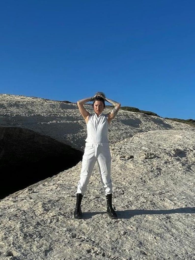 K&ouml;nnte beinhalten: Ein &auml;rmelloser wei&szlig;er Jumpsuit mit Frontrei&szlig;verschluss und Stehkragen, kombiniert mit schwarzen wadenhohen Stiefeln. Der Jumpsuit hat ein k&ouml;rperbetontes Oberteil und eine gerade geschnittene Hose, was einen monochromen Look vor einer Felslandschaft ergibt.