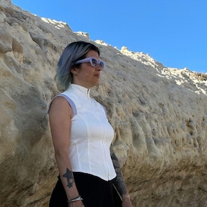 May include: A sleeveless white top with a high collar and a full-length front zipper, featuring light purple trim on the shoulders, worn with black bottoms and light purple-framed sunglasses.