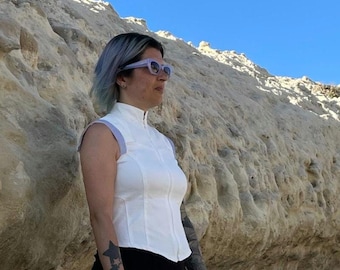 May include: A sleeveless white top with a high collar and a full-length front zipper, featuring light purple trim on the shoulders, worn with black bottoms and light purple-framed sunglasses.