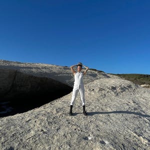 May include: A white sleeveless jumpsuit with a collared neckline and a front zipper closure, paired with black mid-calf lace-up boots. The garment features a fitted bodice and straight-leg trousers, creating a clean and uniform appearance.