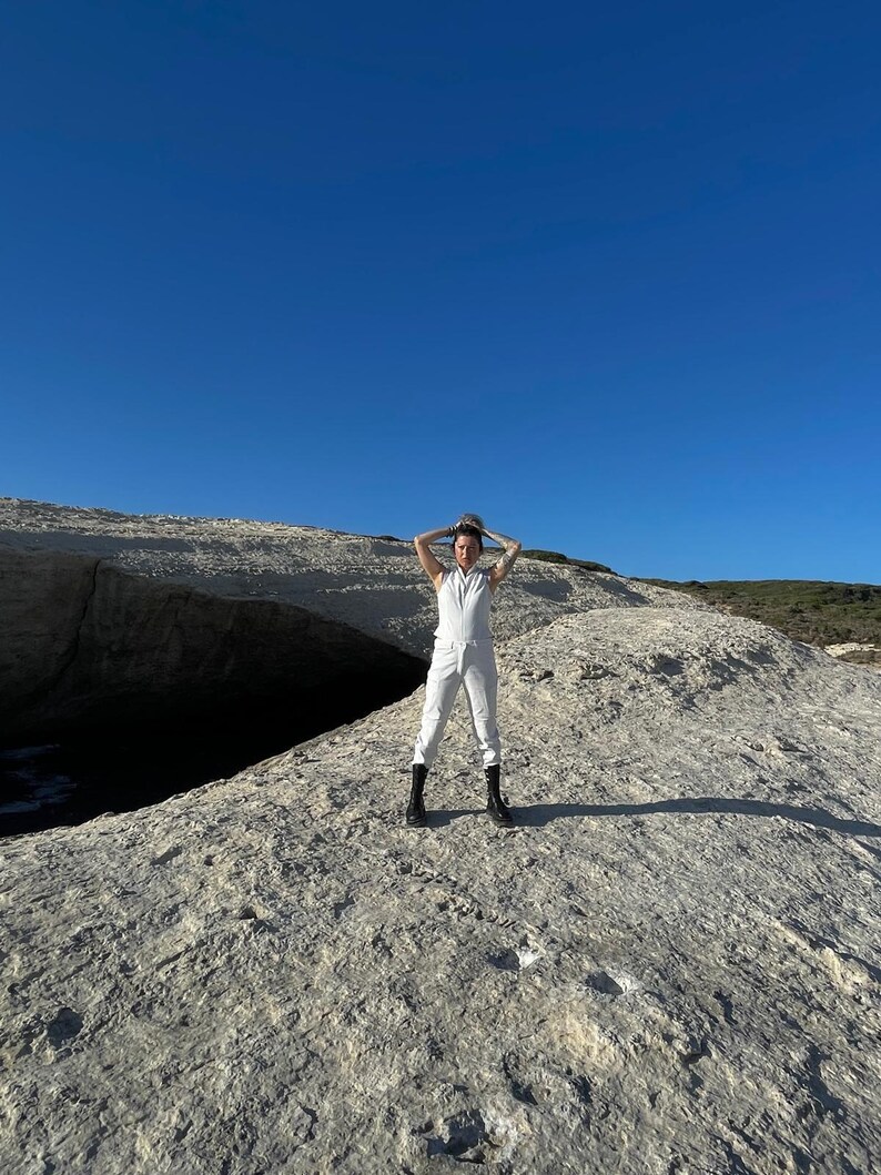 May include: A white sleeveless jumpsuit with a collared neckline and a front zipper closure, paired with black mid-calf lace-up boots. The garment features a fitted bodice and straight-leg trousers, creating a clean and uniform appearance.