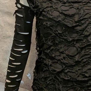 May include: A black long-sleeve top featuring a highly textured, crinkled fabric surface. The sleeve is designed with a series of horizontal cutouts or slashes running down the length of the arm, creating a distressed appearance.