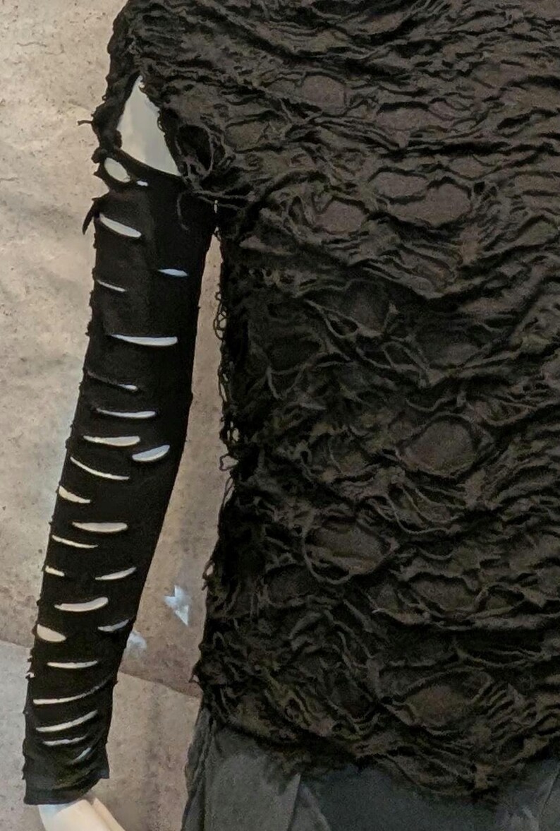 May include: A black long-sleeve top featuring a highly textured, crinkled fabric surface. The sleeve is designed with a series of horizontal cutouts or slashes running down the length of the arm, creating a distressed appearance.