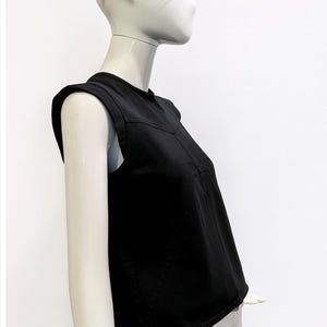 May include: A black sleeveless top with structured shoulders and a V-shaped seam across the chest, worn by a mannequin sporting futuristic translucent blue-tinted visor glasses with black side frames against a plain white background.