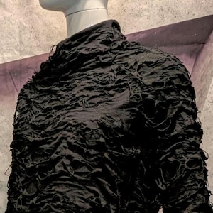 May include: A black long-sleeve top with a high neckline, featuring a heavily distressed, shredded texture across the entire surface. The fabric has numerous irregular holes and frayed edges, creating a layered, uneven appearance on the garment.