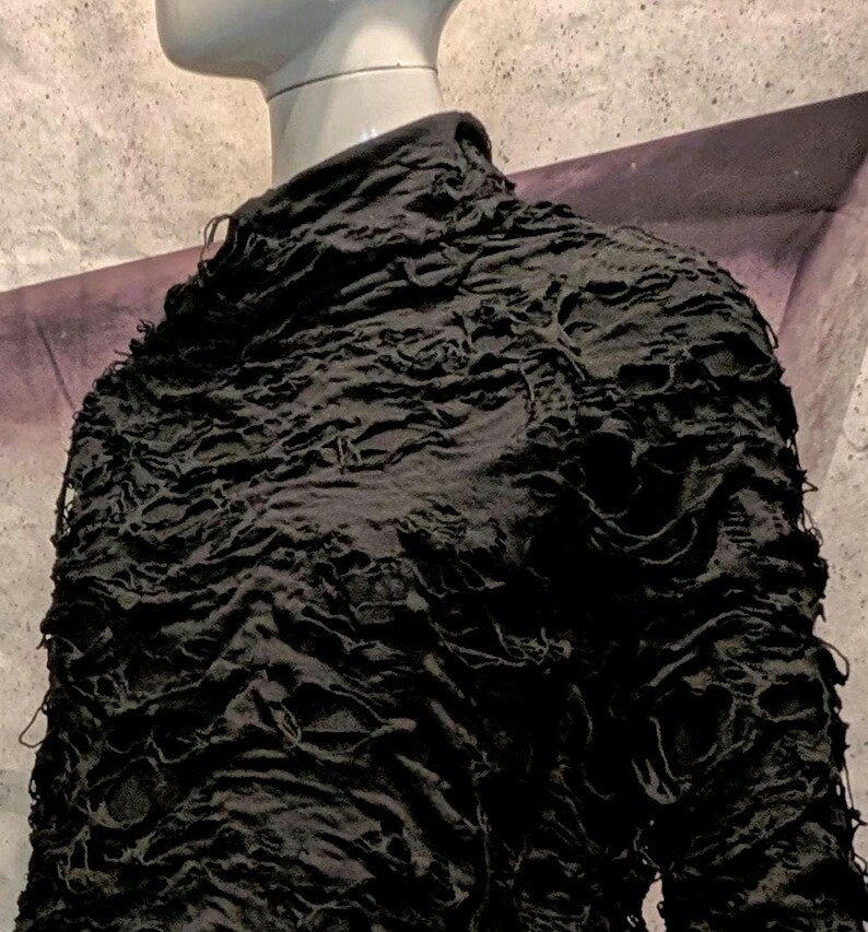May include: A black long-sleeve top with a high neckline, featuring a heavily distressed, shredded texture across the entire surface. The fabric has numerous irregular holes and frayed edges, creating a layered, uneven appearance on the garment.