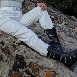 K&ouml;nnte beinhalten: Wei&szlig;e Cargohose mit passendem wei&szlig;em G&uuml;rtel und schwarzen wadenhohen Stiefeln, die mehrere horizontale Riemen auf der Vorderseite aufweisen. Die Stiefel haben eine abgerundete Spitze und eine dicke, dunkle Sohle, die zur hellen Hose kontrastiert.