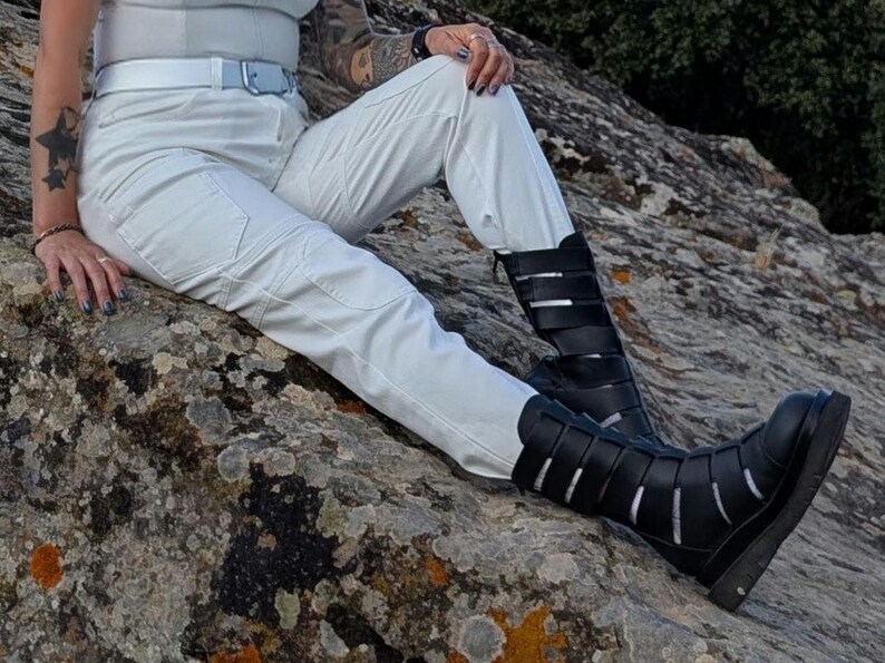 K&ouml;nnte beinhalten: Wei&szlig;e Cargohose mit passendem wei&szlig;em G&uuml;rtel und schwarzen wadenhohen Stiefeln, die mehrere horizontale Riemen auf der Vorderseite aufweisen. Die Stiefel haben eine abgerundete Spitze und eine dicke, dunkle Sohle, die zur hellen Hose kontrastiert.