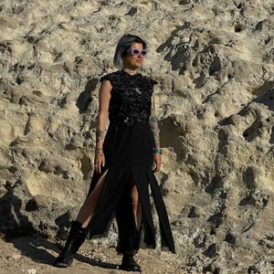 Puede incluir: Un vestido negro sin mangas con un corpi&ntilde;o texturizado y adornado con aplicaciones florales, y una falda larga y fluida con dos aberturas frontales prominentes. La prenda se combina con botas negras con cordones y gafas de sol claras.